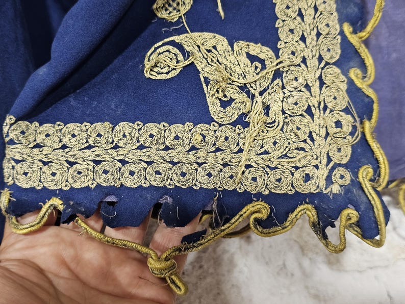 K&ouml;nnte beinhalten: Nahaufnahme eines Abschnitts eines dunkelblauen Stoffes mit aufwendiger goldener Stickerei. Die Stickerei zeigt florale und geometrische Muster entlang der Kanten. Der Stoff hat einen gewellten Rand mit goldenem Besatz.
