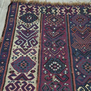 May include: A rectangular woven rug featuring a repeating geometric pattern in shades of deep purple, navy blue, and cream. The textile displays intricate diamond and hooked motifs arranged in vertical bands, finished with a frayed fringe edge.