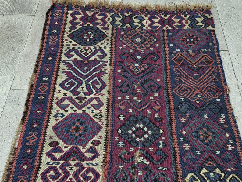 May include: A rectangular woven rug featuring a repeating geometric pattern in shades of deep purple, navy blue, and cream. The textile displays intricate diamond and hooked motifs arranged in vertical bands, finished with a frayed fringe edge.