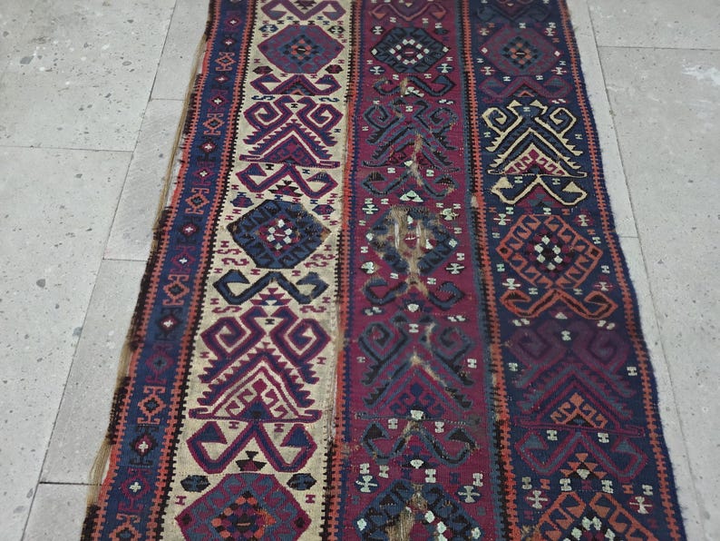 May include: A rectangular woven rug featuring vertical panels with intricate geometric patterns in shades of deep red, navy blue, cream, and dark purple. The textile displays repeating diamond and angular motifs throughout the entire surface area.