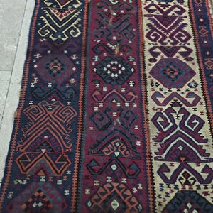 May include: A woven runner rug featuring three vertical panels with intricate geometric patterns in shades of deep blue, burgundy, and cream. The textile displays repeating diamond and angular motifs throughout, with frayed edges along the sides.