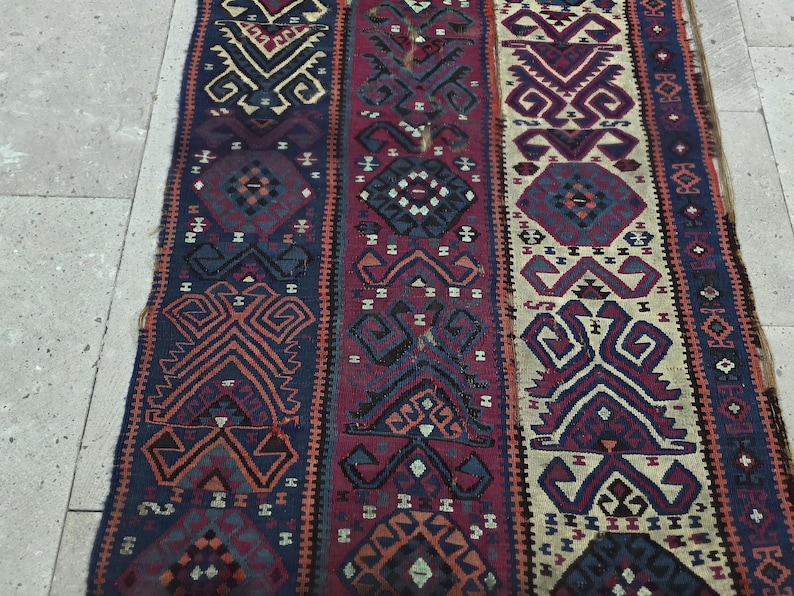 May include: A woven runner rug featuring three vertical panels with intricate geometric patterns in shades of deep blue, burgundy, and cream. The textile displays repeating diamond and angular motifs throughout, with frayed edges along the sides.
