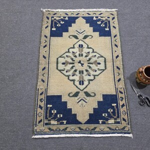 May include: A rectangular area rug featuring a central diamond motif in beige, blue, and green tones. The rug has a deep blue border with geometric patterns and white fringe at both ends. A brown ceramic mug and metal scissors are placed nearby.
