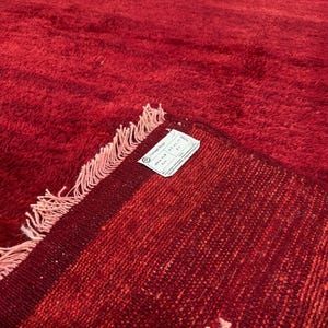 Op de afbeelding: Een dieprood tapijt met een dikke, pluizige poolstructuur en een omgevouwen hoek die de geweven achterkant laat zien. Een wit rechthoekig label is aan de hoek bevestigd, met de tekst Vintage Rugs en de afmetingen 100 x 149 cm.