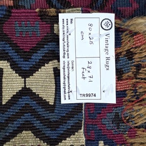 May include: A woven rug featuring a geometric pattern in shades of deep red, blue, and beige. A white label is attached, displaying the text Vintage Rugs, 80x215 cm, 2.8x7.1 feet, and TR9974, along with contact information and web addresses.