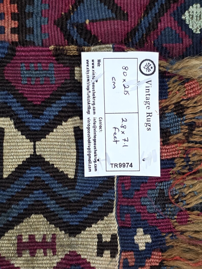 May include: A woven rug featuring a geometric pattern in shades of deep red, blue, and beige. A white label is attached, displaying the text Vintage Rugs, 80x215 cm, 2.8x7.1 feet, and TR9974, along with contact information and web addresses.