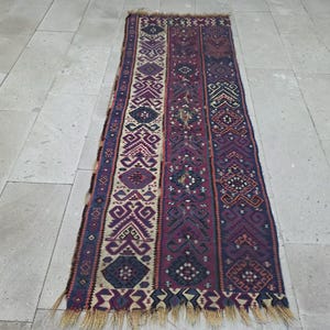 May include: A rectangular woven runner rug featuring a vertical striped pattern in shades of deep purple, navy blue, and cream. The design incorporates intricate geometric motifs and diamond shapes throughout, finished with a fringed edge at both ends.