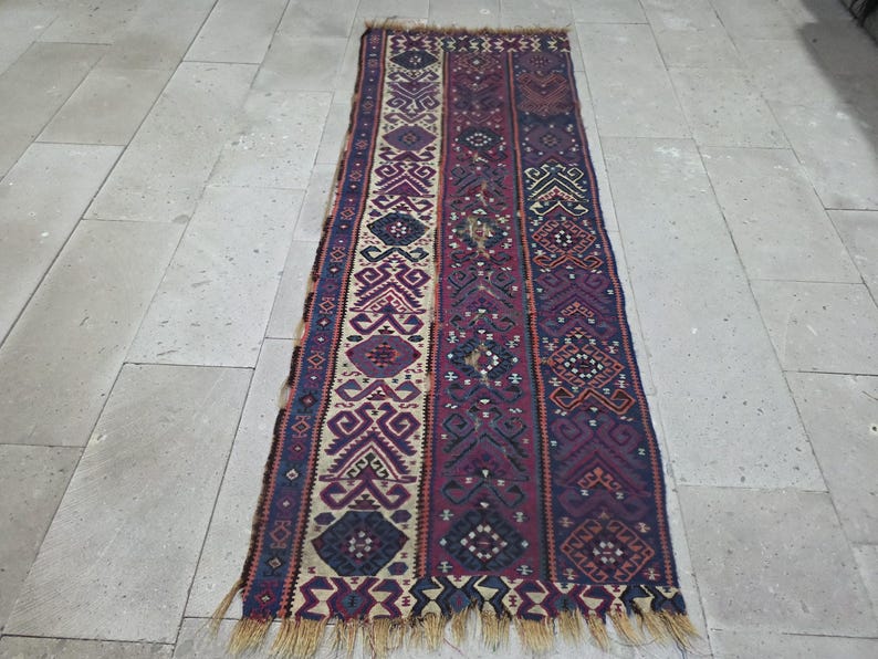 May include: A rectangular woven runner rug featuring a vertical striped pattern in shades of deep purple, navy blue, and cream. The design incorporates intricate geometric motifs and diamond shapes throughout, finished with a fringed edge at both ends.