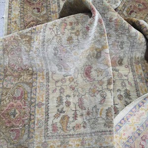 May include: An antique rug with a faded, distressed appearance. The rug features a beige background with intricate floral and geometric patterns in shades of pink, brown, and gold. The edges are bordered with a detailed design.