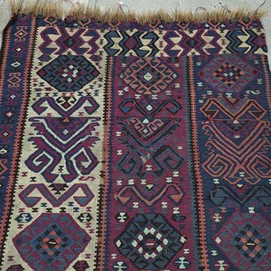 May include: A rectangular woven rug featuring a repeating geometric pattern in shades of deep blue, maroon, and cream. The textile displays intricate symmetrical motifs and stylized shapes throughout, finished with a frayed fringe along the top edge.