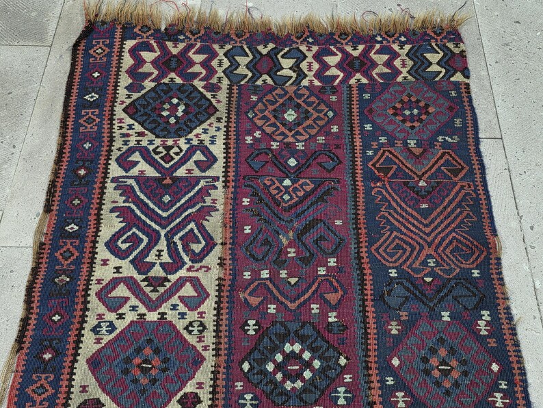 May include: A rectangular woven rug featuring a repeating geometric pattern in shades of deep blue, maroon, and cream. The textile displays intricate symmetrical motifs and stylized shapes throughout, finished with a frayed fringe along the top edge.