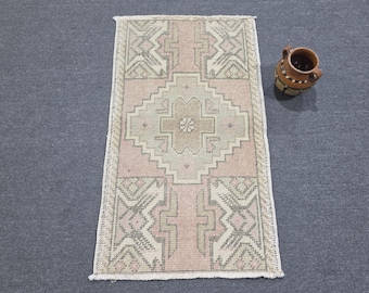 May include: A rectangular area rug featuring a central diamond motif in muted pink and sage green tones with geometric border patterns and short white fringe on the ends, accompanied by a small brown ceramic pitcher with a dark rim.