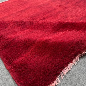 Op de afbeelding: Een dieprood, hoogpolig vloerkleed met een dikke, shaggy textuur. Het kleed is dubbelgevouwen en toont een dicht, pluizig oppervlak en een rechte rand die is afgewerkt met korte, lichtgekleurde franjes langs de onderzijde.