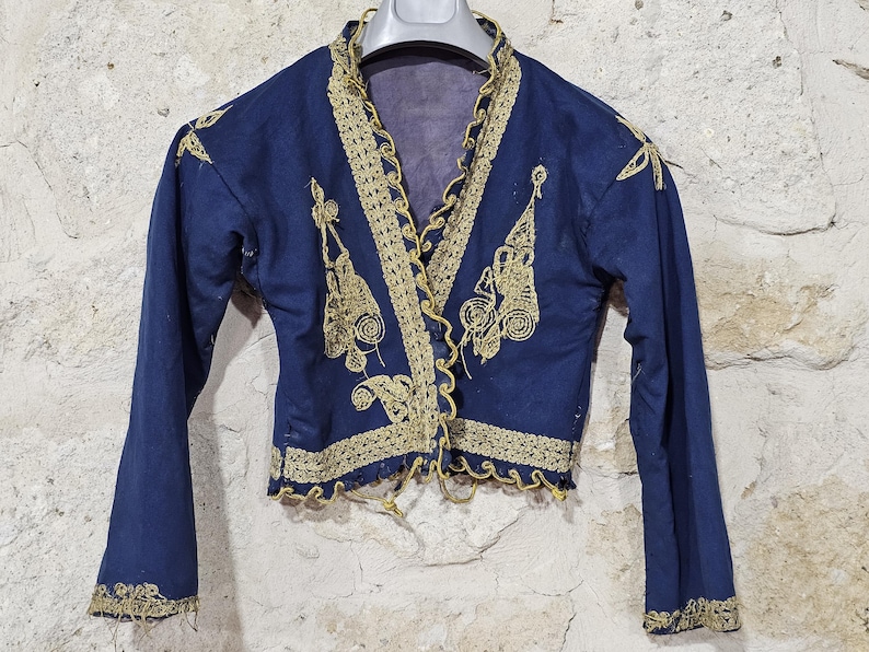 K&ouml;nnte beinhalten: Eine marineblaue Vintage-Jacke mit goldener Stickerei. Die Jacke hat aufwendige goldene Details am Kragen, an den vorderen Kanten und an den Manschetten. Das Design umfasst florale und scrollartige Muster. Die Jacke wird auf einem Kleiderb&uuml;gel pr&auml;sentiert.