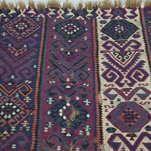 May include: A woven rug featuring vertical panels in deep blue, burgundy, and cream. The textile displays intricate geometric patterns, including diamonds and stylized motifs, with a fringed edge at the top. The weave shows visible signs of wear.