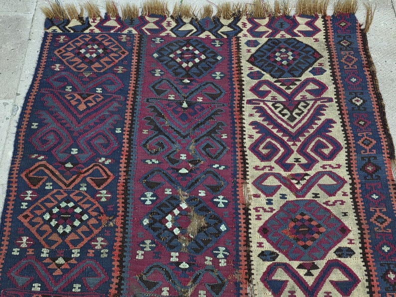 May include: A woven rug featuring vertical panels in deep blue, burgundy, and cream. The textile displays intricate geometric patterns, including diamonds and stylized motifs, with a fringed edge at the top. The weave shows visible signs of wear.