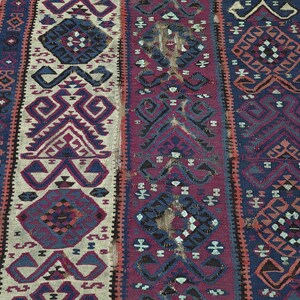 May include: A woven rug featuring three vertical panels with geometric patterns in shades of deep blue, burgundy, and cream. The design includes repeating diamond shapes and angular motifs throughout the textile, showcasing a textured, flat-weave surface.
