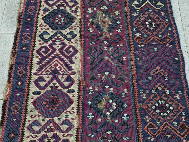 May include: A woven rug featuring three vertical panels with geometric patterns in shades of deep blue, burgundy, and cream. The design includes repeating diamond shapes and angular motifs throughout the textile, showcasing a textured, flat-weave surface.