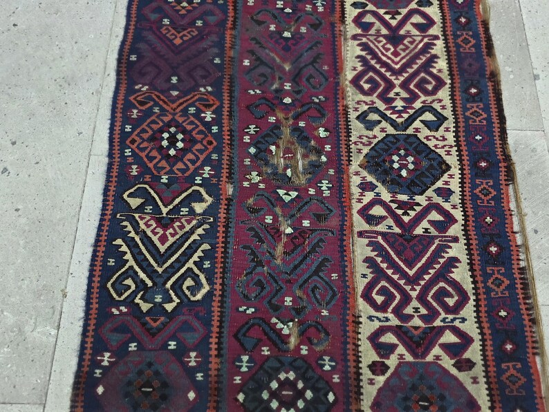 May include: A rectangular woven textile runner featuring three vertical panels with intricate geometric patterns in shades of deep blue, dark red, and cream. The design includes repeating diamond and hooked motifs with visible frayed edges.
