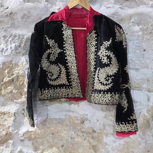 以下が含まれることがあります： 金色の刺繍と赤い裏地が付いた黒いベルベットのジャケットが、木製のハンガーに掛けられています。 ジャケットは、縁、袖口、フロントパネルに金色の装飾が施されています。 ジャケットは、質感のある石壁を背景に展示されています。