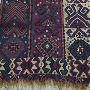 May include: A woven rug featuring vertical panels with geometric patterns in shades of deep blue, burgundy, and cream. The design includes repeating diamond and angular motifs throughout the fabric, finished with a frayed fringe along the bottom edge.
