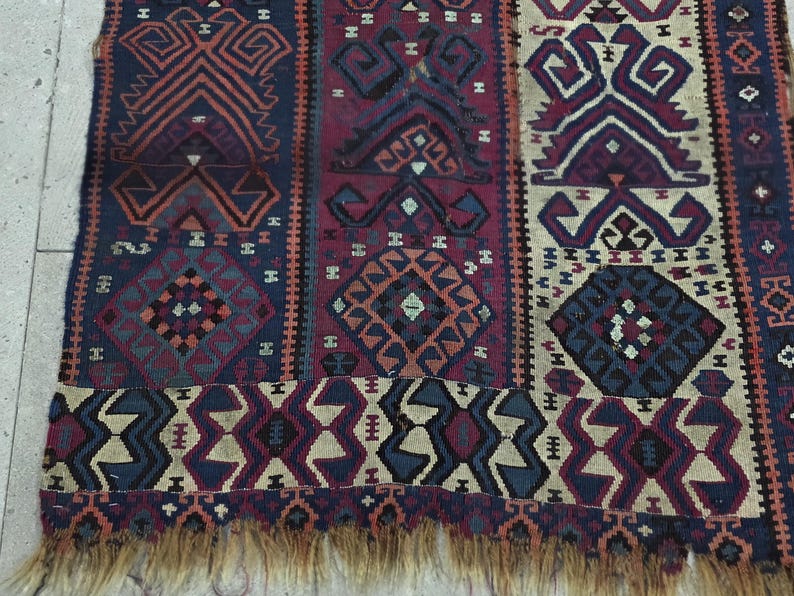 May include: A woven rug featuring vertical panels with geometric patterns in shades of deep blue, burgundy, and cream. The design includes repeating diamond and angular motifs throughout the fabric, finished with a frayed fringe along the bottom edge.