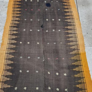 May include: A black and gold woven rug with a geometric pattern. The rug has a fringe border and is made of natural fibres.