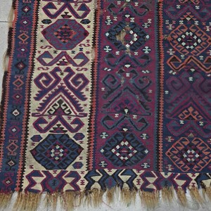 May include: A woven textile rug featuring vertical panels with geometric patterns in shades of deep red, navy blue, and cream. The design includes repeating diamond and angular motifs with a frayed, light brown fringe along the bottom edge.