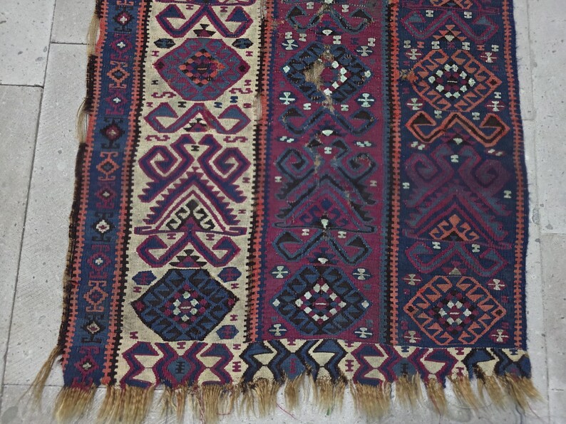 May include: A woven textile rug featuring vertical panels with geometric patterns in shades of deep red, navy blue, and cream. The design includes repeating diamond and angular motifs with a frayed, light brown fringe along the bottom edge.