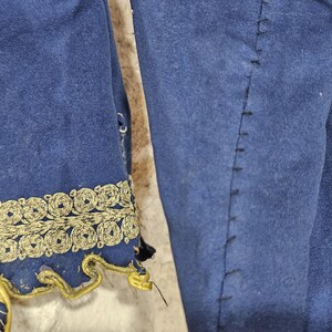 K&ouml;nnte beinhalten: Nahaufnahme eines blauen Kleidungsst&uuml;cks mit goldener Stickerei. Die goldene Verzierung verziert den &Auml;rmelrand mit einem sich wiederholenden kreisf&ouml;rmigen Design. Der Stoff hat eine weiche, Wildleder-&auml;hnliche Textur. Kost&uuml;mkleidung.