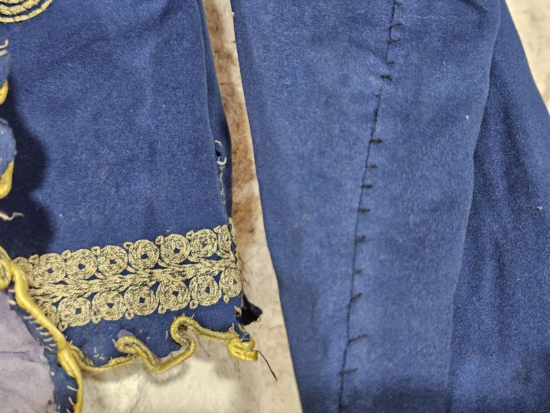 K&ouml;nnte beinhalten: Nahaufnahme eines blauen Kleidungsst&uuml;cks mit goldener Stickerei. Die goldene Verzierung verziert den &Auml;rmelrand mit einem sich wiederholenden kreisf&ouml;rmigen Design. Der Stoff hat eine weiche, Wildleder-&auml;hnliche Textur. Kost&uuml;mkleidung.
