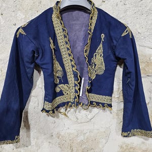 K&ouml;nnte beinhalten: Dunkelblaue Jacke mit goldener Stickerei. Die Jacke hat verzierte goldene Details am Kragen, an den Manschetten und an den vorderen Kanten. Die Stickerei umfasst Blumen- und Spiralmotive. Die Jacke wird vor einer strukturierten, hellfarbigen Wand pr&auml;sentiert.