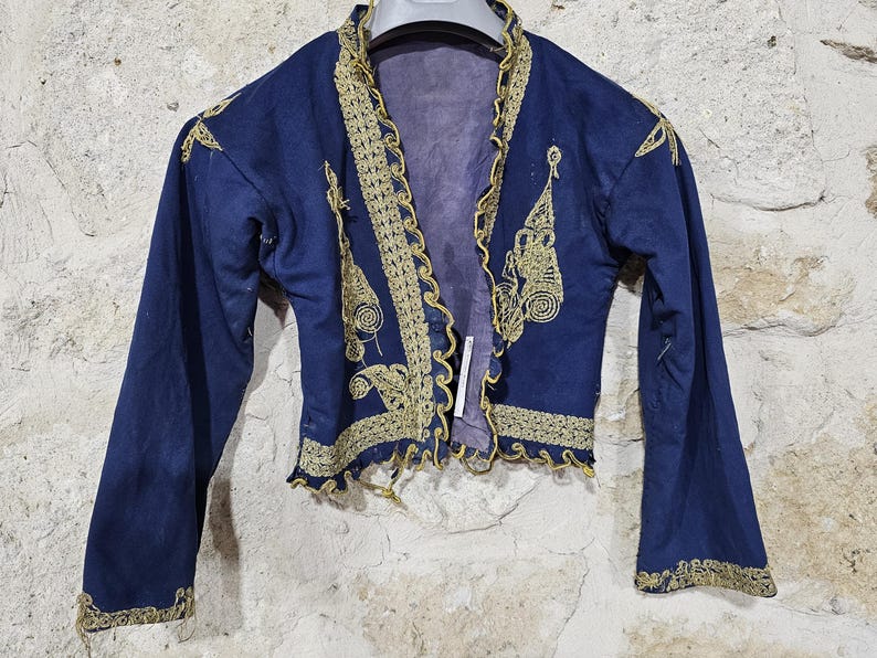 K&ouml;nnte beinhalten: Dunkelblaue Jacke mit goldener Stickerei. Die Jacke hat verzierte goldene Details am Kragen, an den Manschetten und an den vorderen Kanten. Die Stickerei umfasst Blumen- und Spiralmotive. Die Jacke wird vor einer strukturierten, hellfarbigen Wand pr&auml;sentiert.