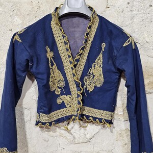 K&ouml;nnte beinhalten: Marineblaue Jacke mit goldener Stickerei. Die Jacke hat aufwendige goldene Verzierungen am Kragen, an der Vorderseite und an den Manschetten. Das Design beinhaltet florale und geometrische Muster. Die Jacke wird auf einem Kleiderb&uuml;gel vor einer Steinmauer pr&auml;sentiert.