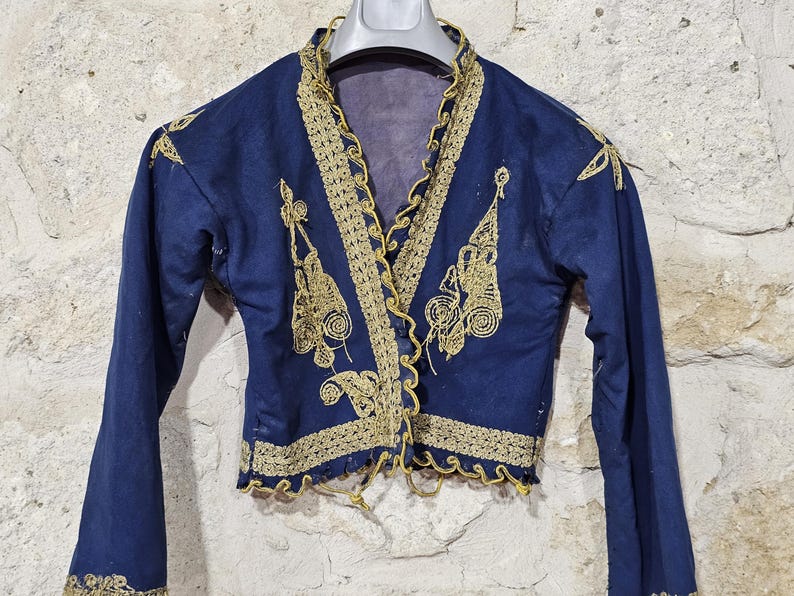 K&ouml;nnte beinhalten: Marineblaue Jacke mit goldener Stickerei. Die Jacke hat aufwendige goldene Verzierungen am Kragen, an der Vorderseite und an den Manschetten. Das Design beinhaltet florale und geometrische Muster. Die Jacke wird auf einem Kleiderb&uuml;gel vor einer Steinmauer pr&auml;sentiert.