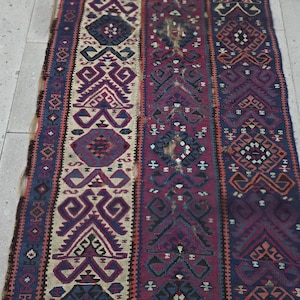 May include: A rectangular woven rug featuring vertical panels with intricate geometric patterns in shades of deep red, navy blue, cream, and dark purple. The design consists of repeating diamond and angular motifs arranged in symmetrical columns.