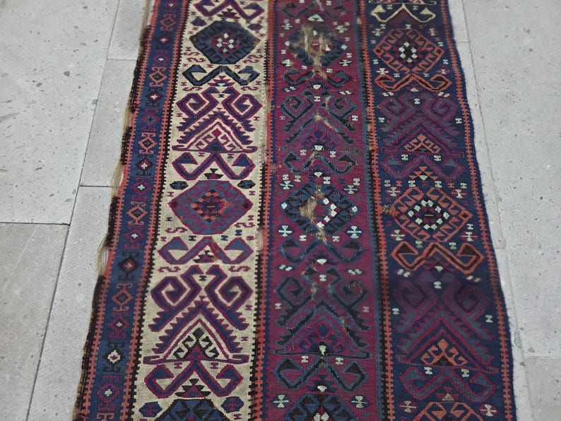 May include: A rectangular woven rug featuring vertical panels with intricate geometric patterns in shades of deep red, navy blue, cream, and dark purple. The design consists of repeating diamond and angular motifs arranged in symmetrical columns.
