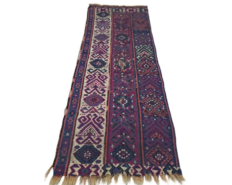 May include: A long, rectangular runner rug featuring a series of vertical panels with intricate geometric patterns. The color palette includes deep shades of purple, magenta, navy blue, and cream. The rug is finished with a light tan fringe at the ends.