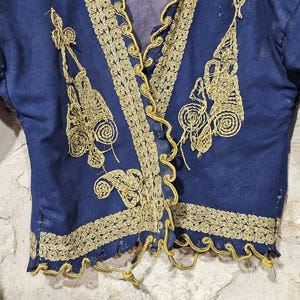 K&ouml;nnte beinhalten: Dunkelblaue Jacke mit goldener Stickerei. Die Jacke hat aufwendige Goldfaden-Designs entlang der Kanten, des Halsausschnitts und der Frontplatten. Die Stickerei umfasst Blumen- und Scrollmuster. Die Jacke hat lange &Auml;rmel und einen V-Ausschnitt.