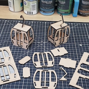 Peut inclure: Un ensemble de kits de modèles de téléphériques en bois. Les kits comprennent des pièces de bois prédécoupées et deux téléphériques assemblés. Les kits sont disposés sur un tapis de coupe noir.