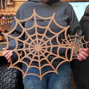 May include: A person holding two wooden spiderweb decorations. The larger web is approximately 24 inches in diameter, and the smaller one is about 12 inches. The webs are made of light brown wood and are suitable for Halloween decor.