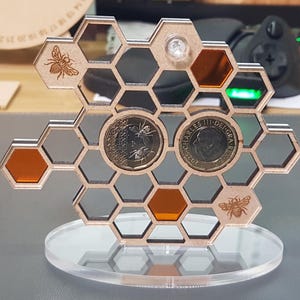 May include: A wooden honeycomb-shaped coin display with engraved bee designs and mirrored and orange-tinted hexagonal cells. The display holds two coins and sits on a clear acrylic base.