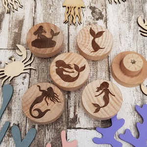 Könnte beinhalten: Holzbastelsortiment mit verschiedenen Meeresmotiven, darunter Meerjungfrauen, Krabben und Quallen. Die Formen sind aus Naturholz geschnitten und haben einen rustikalen, unfertigen Look.