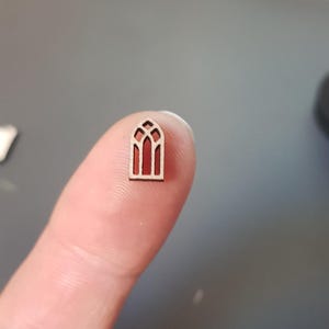 May include: A small wooden cutout of a stained glass window with a red interior. The window is arched and has a simple design.