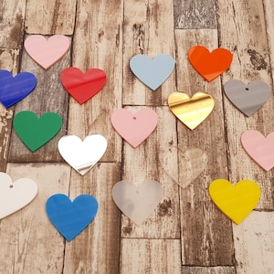 May include: Assortment of 15 heart-shaped acrylic blanks in various colours, including red, blue, green, pink, yellow, gold, and silver. The blanks are arranged on a wooden surface.