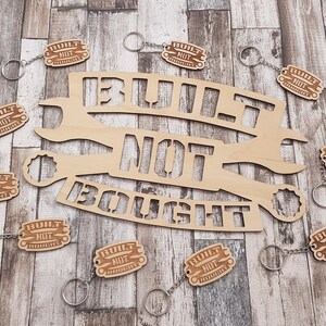 May include: Wooden keychains and a large wooden sign with the phrase "BUILT NOT BOUGHT" cut out. The keychains have a small tag with the same phrase. The items are light brown and sit on a wood-grain surface.