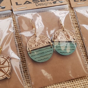 May include: Three pairs of wooden earrings with different designs. The first pair features a compass design, the second pair features a mountain and lake design, and the third pair features a compass design. The earrings are packaged in clear plastic bags with a brown paper backing.