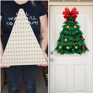 May include: A wooden triangle with holes for inserting items, like a Christmas tree, for crafting a wreath. The triangle is being held by a person. A green and gold wreath with red ornaments and a red bow is hanging on a white door.