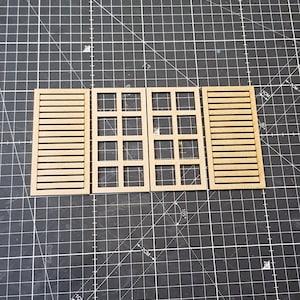 May include: Wooden miniature architectural elements, including window frames and shutters, are arranged on a black cutting mat with grid lines. The pieces are made of light-colored wood and are likely for a model or craft project.