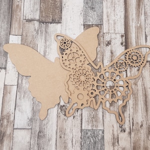 May include: A wooden butterfly cutout with intricate gear designs on one wing. The butterfly is a light brown color and is set against a background of wood planks.